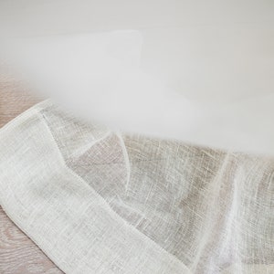 Puede incluir: Primer plano de una tela de lino blanco roto doblada, con un tejido texturizado. La tela tiene un color natural sutil y una ca&iacute;da suave y fluida. Los bordes est&aacute;n cuidadosamente acabados, lo que sugiere un textil de alta calidad. El fondo es una superficie de madera clara.