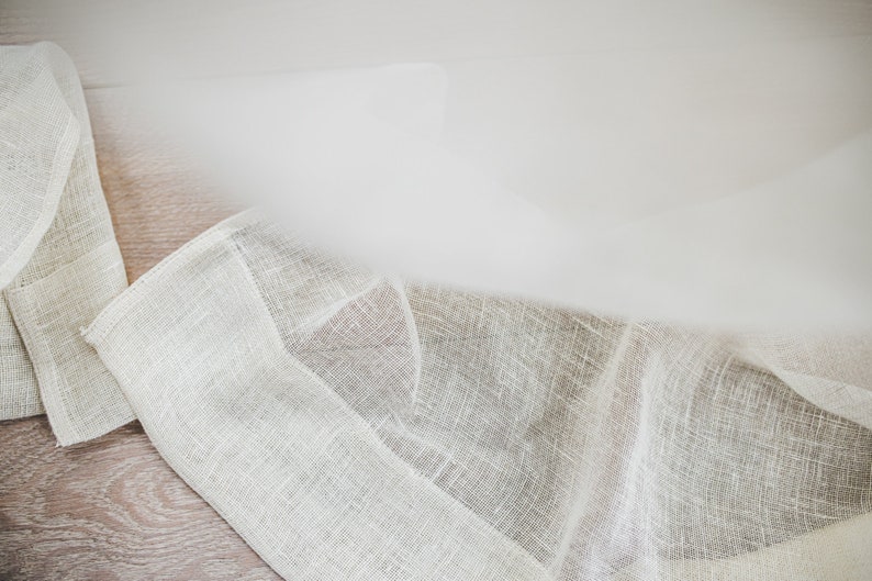 Puede incluir: Primer plano de una tela de lino blanco roto doblada, con un tejido texturizado. La tela tiene un color natural sutil y una ca&iacute;da suave y fluida. Los bordes est&aacute;n cuidadosamente acabados, lo que sugiere un textil de alta calidad. El fondo es una superficie de madera clara.
