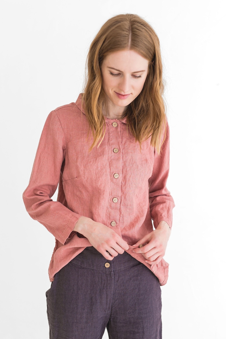 May include: A woman wearing a pink linen shirt with a collar and button fastening. She is also wearing dark purple linen trousers with a button fastening.