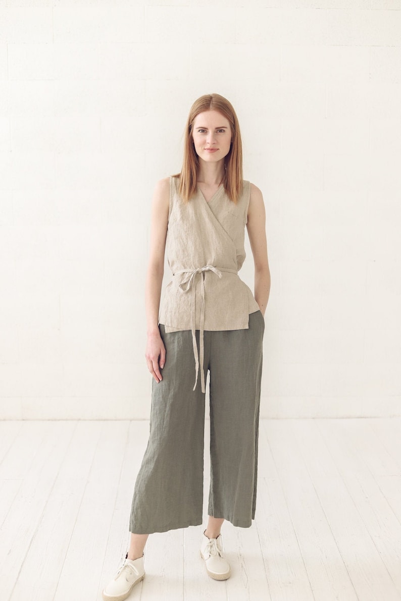 May include: A woman wearing a beige linen wrap top with a matching tie belt and olive green linen wide-leg pants. She is standing on a white wood floor.