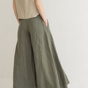 May include: A pair of wide-leg linen pants in a muted olive green color. The pants have a high waist and a flowing, relaxed fit. The model is wearing the pants with a light brown linen top and brown sandals.