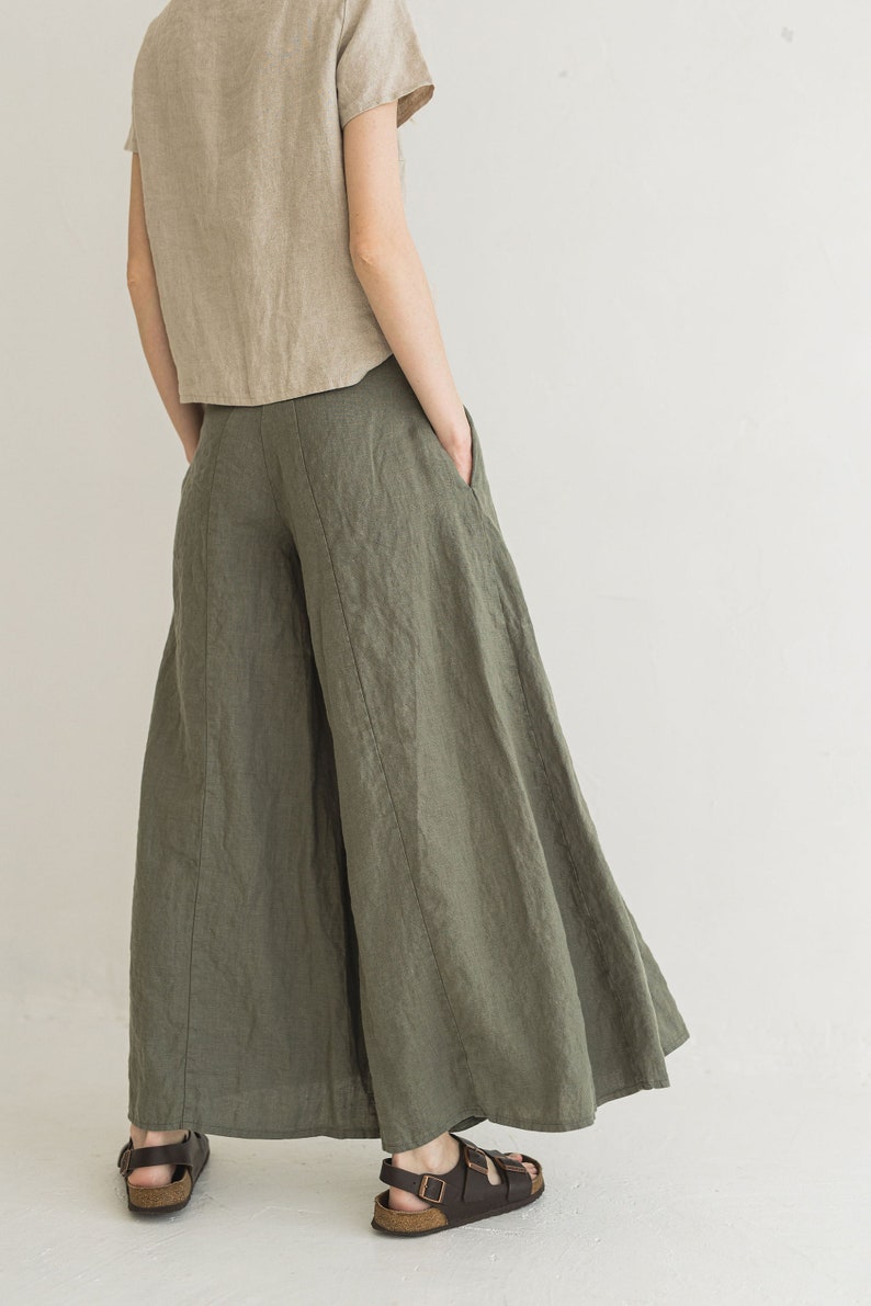 May include: A pair of wide-leg linen pants in a muted olive green color. The pants have a high waist and a flowing, relaxed fit. The model is wearing the pants with a light brown linen top and brown sandals.
