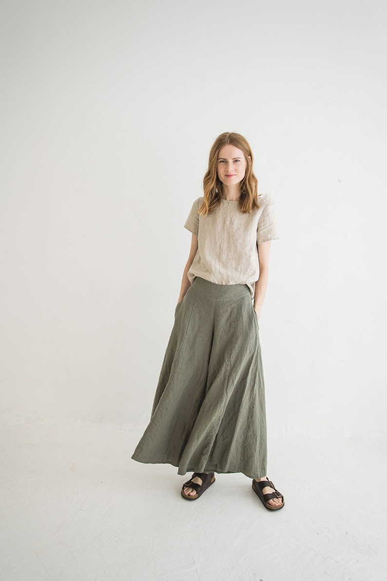 May include: A woman wearing a light beige linen top and wide-legged, olive green linen pants. She is standing in front of a white background and has her hands in her pockets.