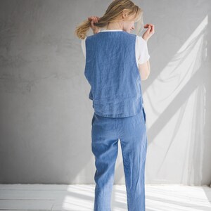 May include: A woman wearing a blue linen jumpsuit with a white sleeveless top. The jumpsuit has a wide leg and a relaxed fit.
