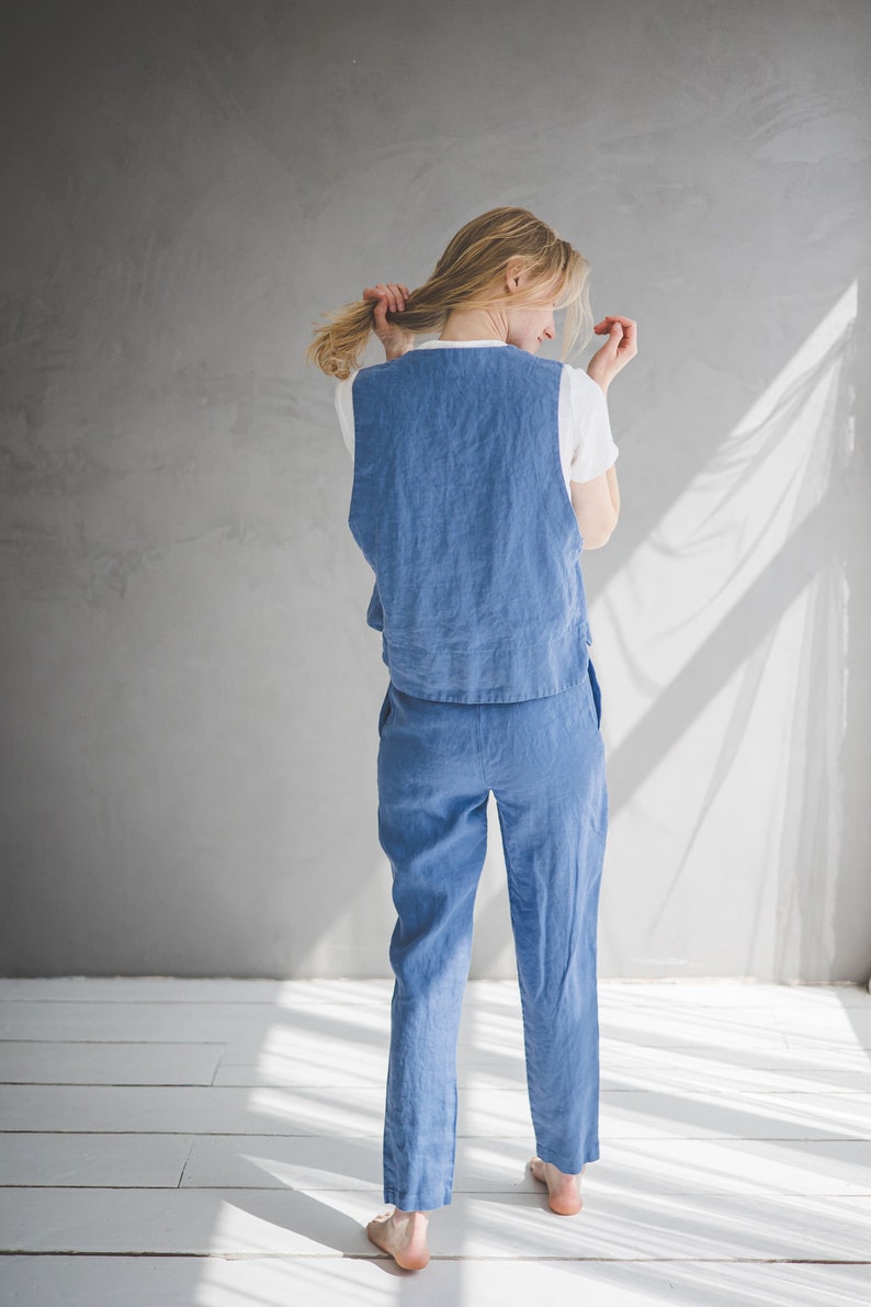 May include: A woman wearing a blue linen jumpsuit with a white sleeveless top. The jumpsuit has a wide leg and a relaxed fit.