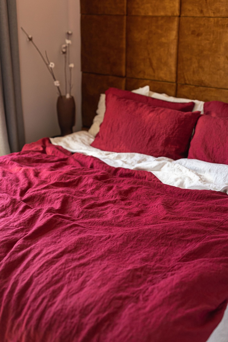 May include: A red linen duvet cover with a white linen sheet underneath. The duvet cover is wrinkled and has a textured surface. The bed is against a brown headboard.