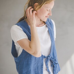 May include: A woman wearing a light blue linen vest with a white t-shirt underneath. The vest has a tie at the waist and the woman is wearing matching light blue pants.