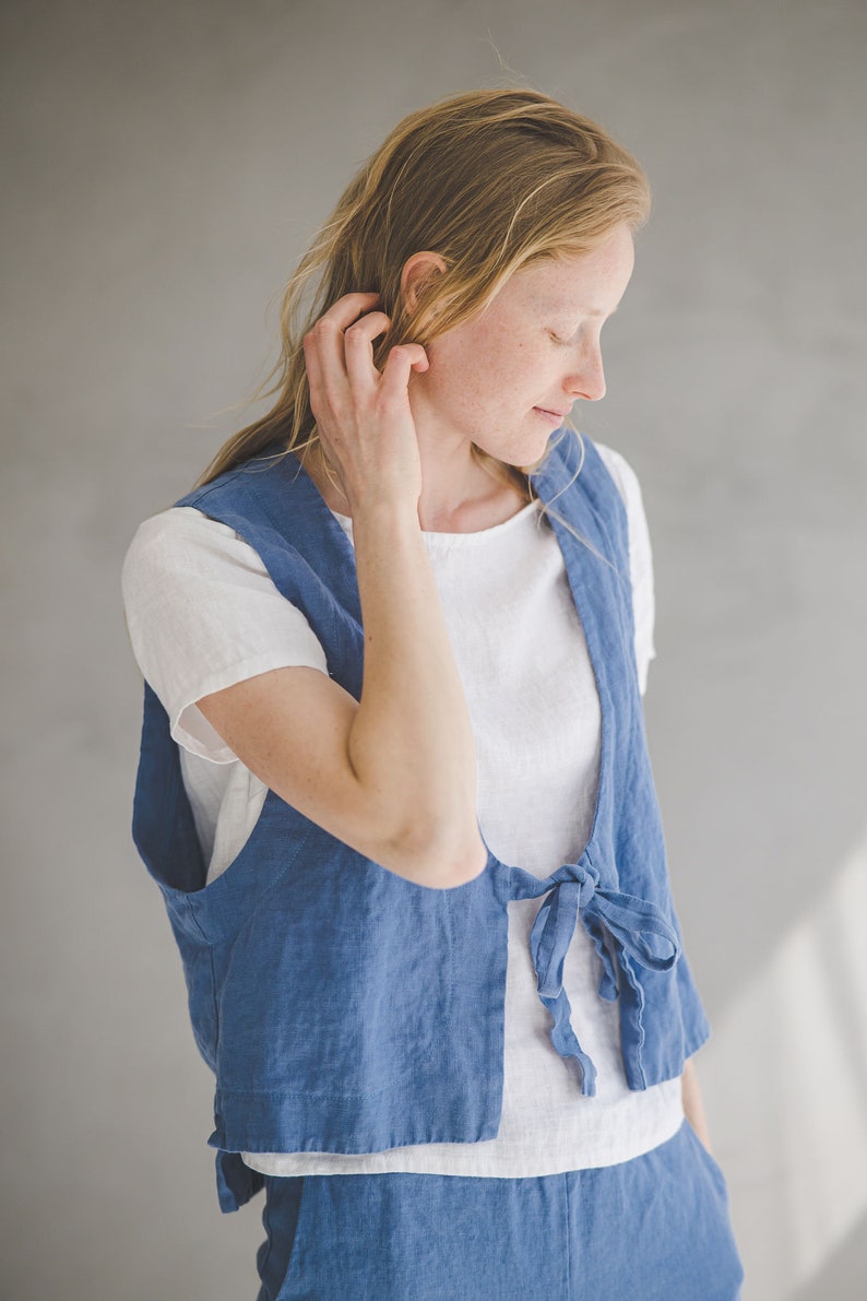 May include: A woman wearing a light blue linen vest with a white t-shirt underneath. The vest has a tie at the waist and the woman is wearing matching light blue pants.