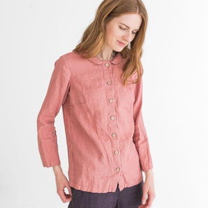 May include: A woman wearing a long-sleeved, pink linen shirt with a peter pan collar and a button-up front. She is also wearing dark grey linen trousers.
