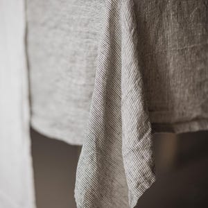 May include: Close-up of a gray and white striped linen tablecloth draped over a table. The fabric has a textured appearance with vertical stripes. The tablecloth is made of a natural material and has a casual, rustic aesthetic.