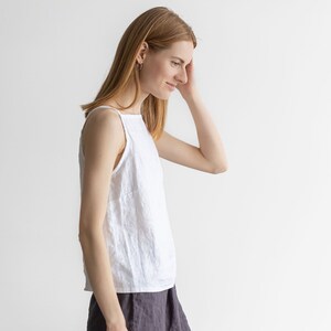 May include: A woman wearing a white linen tank top and a dark gray linen skirt. She is standing in front of a white wall.