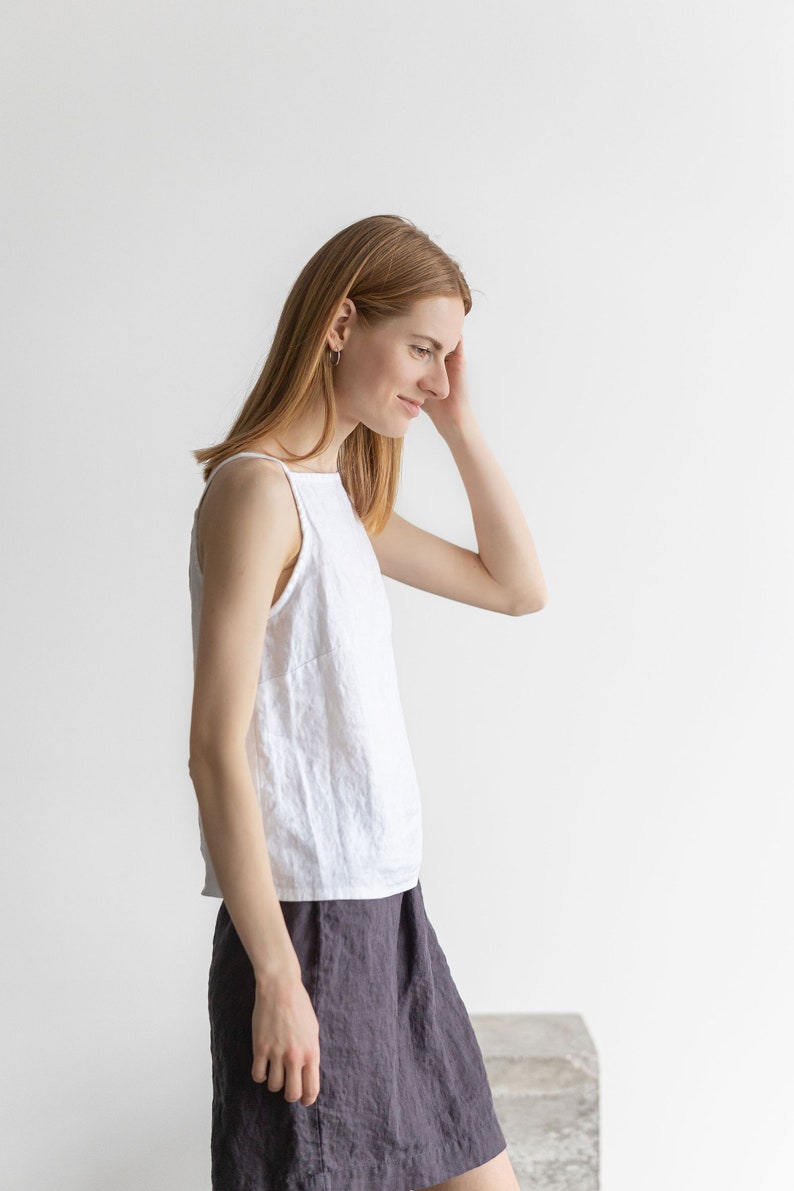 May include: A woman wearing a white linen tank top and a dark gray linen skirt. She is standing in front of a white wall.