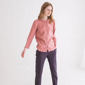 May include: A woman wearing a pink linen shirt with a peter pan collar and long sleeves, paired with dark grey linen trousers and white trainers.