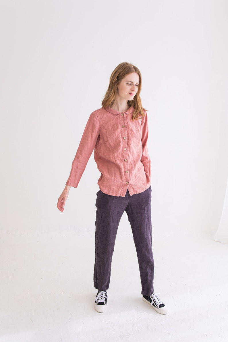 May include: A woman wearing a pink linen shirt with a peter pan collar and long sleeves, paired with dark grey linen trousers and white trainers.