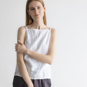 May include: A woman wearing a white linen tank top and a dark gray linen skirt. She is standing in front of a white wall.
