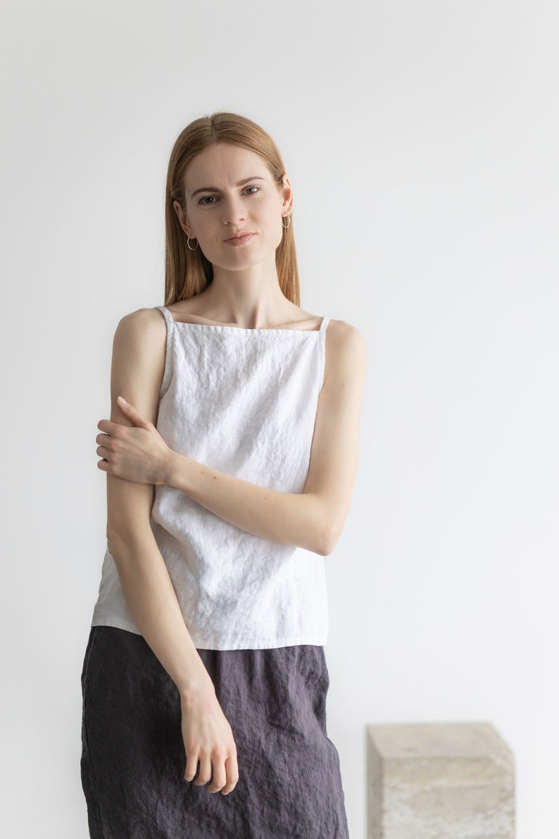 May include: A woman wearing a white linen tank top and a dark gray linen skirt. She is standing in front of a white wall.