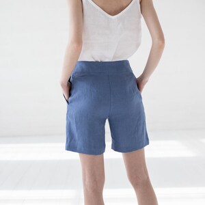 May include: A woman wearing a white linen tank top and blue linen shorts. The shorts have a high waist and pockets.