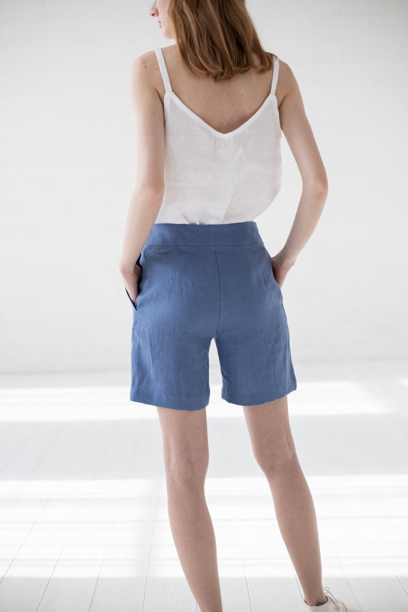 May include: A woman wearing a white linen tank top and blue linen shorts. The shorts have a high waist and pockets.