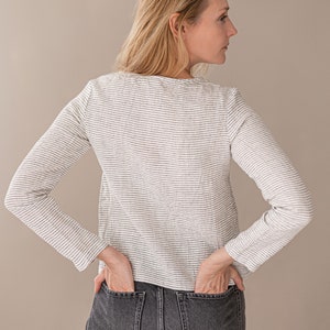 May include: A long-sleeved shirt featuring a white base with thin, horizontal black stripes and a round neckline, paired with dark gray denim jeans that have two visible back pockets. The shirt has a straight hemline and a fitted silhouette.