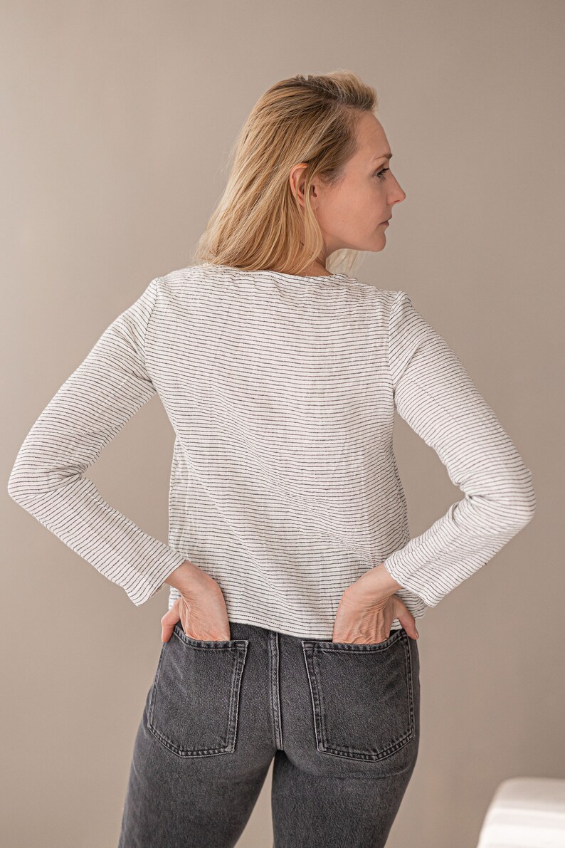 May include: A long-sleeved shirt featuring a white base with thin, horizontal black stripes and a round neckline, paired with dark gray denim jeans that have two visible back pockets. The shirt has a straight hemline and a fitted silhouette.