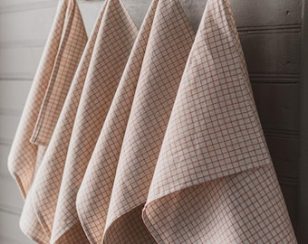 May include: Four cream-colored kitchen towels with a red grid pattern hang from wooden pegs on a wooden board. The towels are folded and draped, showcasing the pattern. The background is a gray, paneled wall.