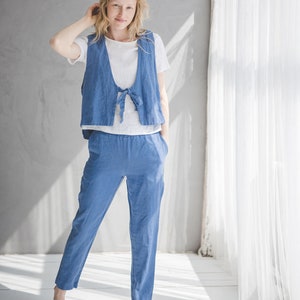 May include: A woman wearing a light blue linen jumpsuit with a tie at the waist. The jumpsuit has a vest-like top and pants. She is standing in a room with white walls and a white curtain in the background.