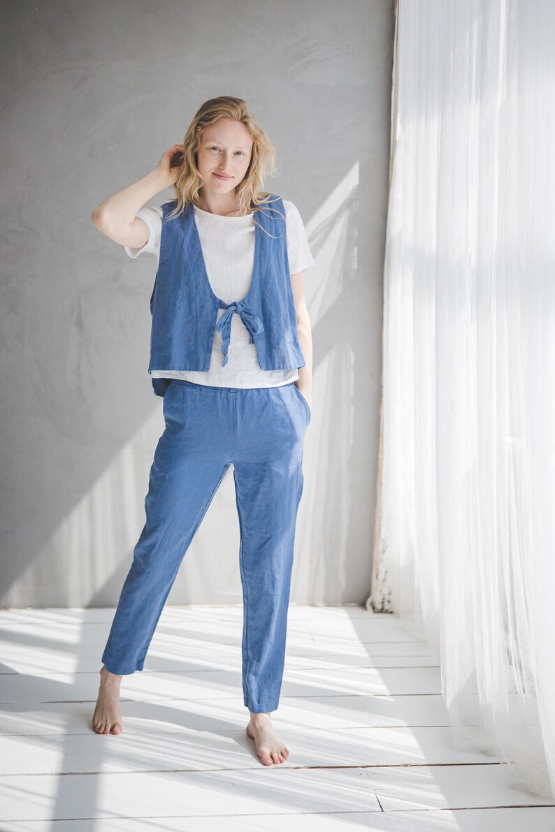 May include: A woman wearing a light blue linen jumpsuit with a tie at the waist. The jumpsuit has a vest-like top and pants. She is standing in a room with white walls and a white curtain in the background.