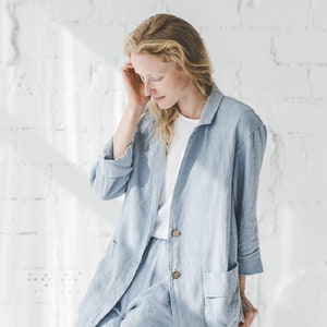 May include: A woman wearing a light blue linen pantsuit with a white t-shirt underneath. She is sitting on a black stool in front of a white brick wall.