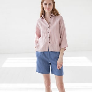 May include: A woman wearing a pink linen shirt with a button-down front and blue linen shorts.