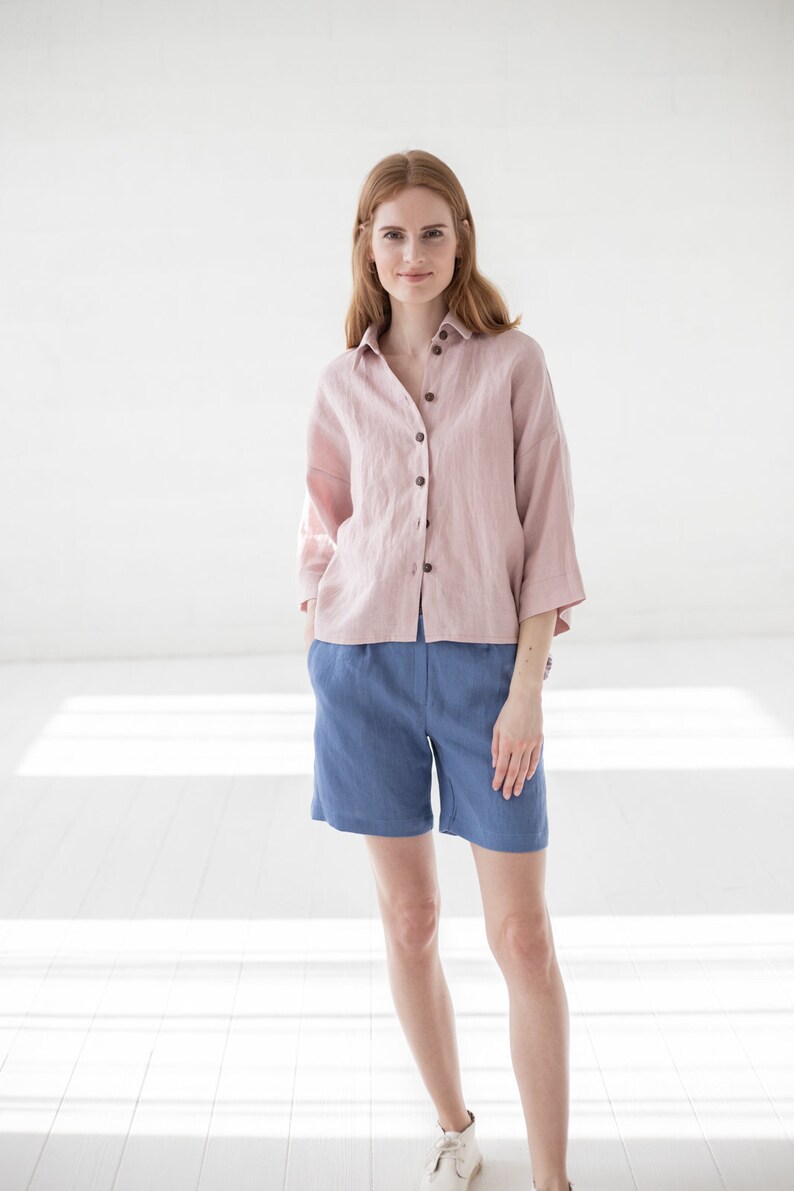 May include: A woman wearing a pink linen shirt with a button-down front and blue linen shorts.