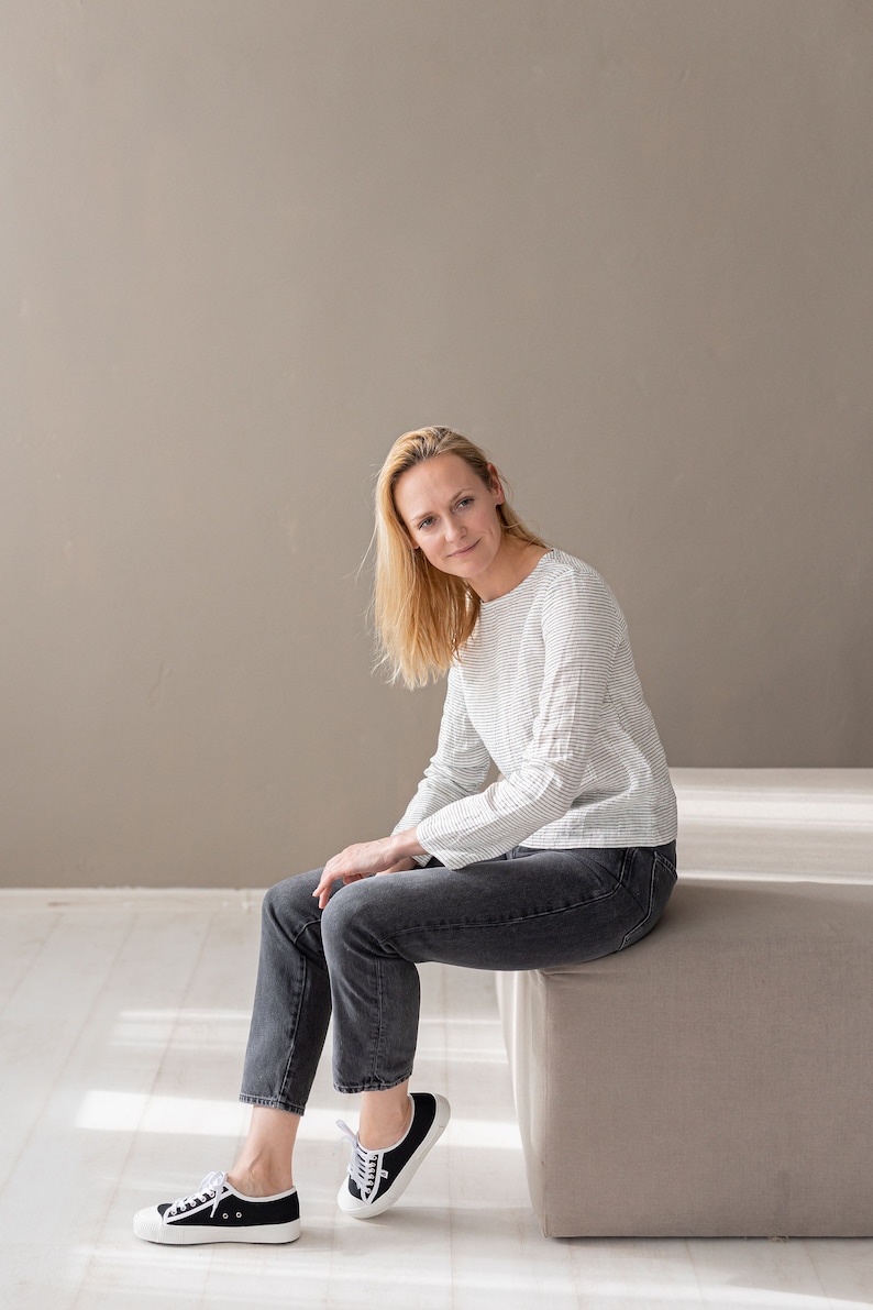 May include: A long-sleeved top featuring a white and black horizontal pinstripe pattern, paired with dark gray denim jeans and black low-top sneakers with white laces and white rubber soles. The top has a round neckline and a straight hemline.