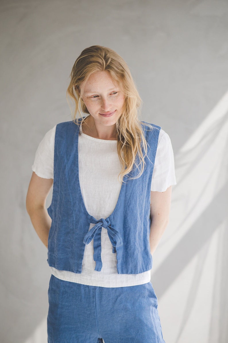 May include: A woman wearing a blue linen vest with a tie closure over a white linen t-shirt and matching blue linen pants.