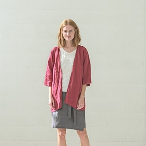 Könnte beinhalten: Eine Frau trägt eine rote Kimono-Jacke aus Leinen, ein weißes V-Ausschnitt-Oberteil und einen grauen Wickelrock. Dazu weiße Schuhe. Der Hintergrund ist eine weiße Backsteinmauer.