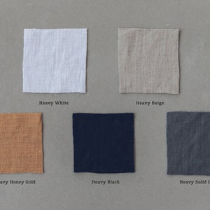 Op de afbeelding: Zes stofstalen in verschillende kleuren: wit, beige, honinggoud, zwart en grijs. Elke staal is gelabeld met de kleur en "Heavy" wat het gewicht van de stof aangeeft.