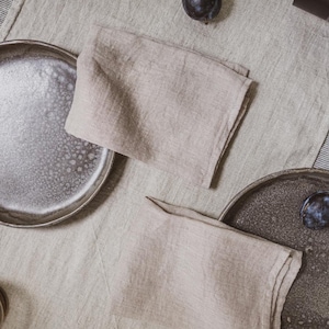 May include: A still-life image of a table setting with two plates, two folded beige linen napkins, and two plums. The plates have a speckled, dark gray finish. The table is covered with a beige linen tablecloth.
