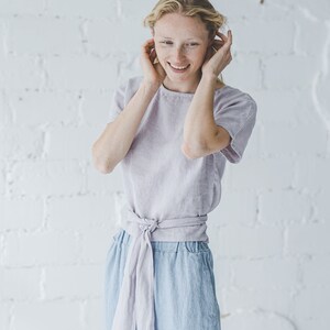 以下が含まれることがあります： 女性は、ウエストに合わせた生地で結ばれた薄い紫色のリネン素材のトップスを着ています。彼女は青いリネンパンツを履いていて、温かい笑顔をしています。