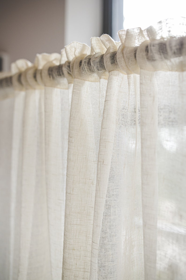 Puede incluir: Una cortina semitransparente de color blanco roto con una textura tejida visible, que presenta un encabezado superior con volantes que se desliza directamente sobre una barra de cortina oscura. La tela ligera cuelga en pliegues verticales.