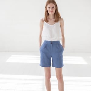 May include: A woman wearing a white linen tank top and blue linen shorts. She is standing in a white room with natural light.