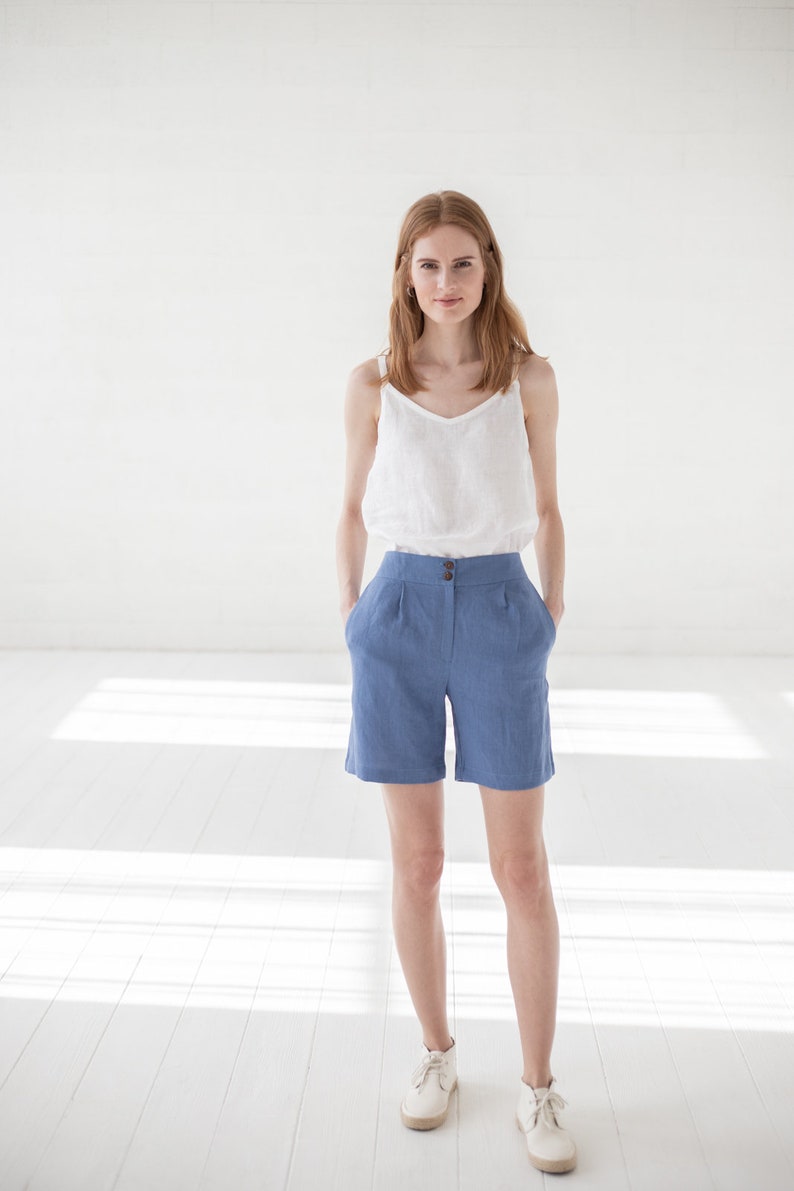 May include: A woman wearing a white linen tank top and blue linen shorts. She is standing in a white room with natural light.