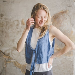 May include: A woman wearing a blue linen vest with a white top and a gray linen skirt. The vest has a tie at the front and the woman is smiling.