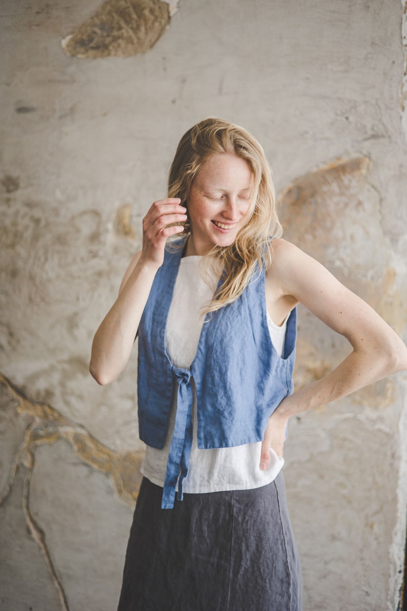 May include: A woman wearing a blue linen vest with a white top and a gray linen skirt. The vest has a tie at the front and the woman is smiling.