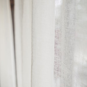 Puede incluir: Un panel de cortina de color blanco roto semitransparente con una textura tejida visible. La tela es ligera y deja pasar la luz a trav&eacute;s del tejido. La cortina cuelga en pliegues verticales, mostrando un patr&oacute;n de tejido consistente.