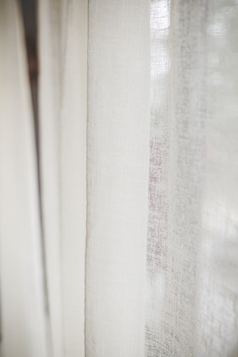 Puede incluir: Un panel de cortina de color blanco roto semitransparente con una textura tejida visible. La tela es ligera y deja pasar la luz a trav&eacute;s del tejido. La cortina cuelga en pliegues verticales, mostrando un patr&oacute;n de tejido consistente.