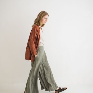 May include: A woman wearing a rust-colored linen jacket, a white linen shirt, and wide-legged green linen pants. She is walking towards the camera, wearing brown sandals.