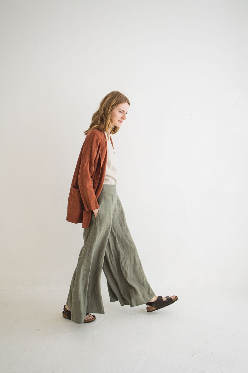May include: A woman wearing a rust-colored linen jacket, a white linen shirt, and wide-legged green linen pants. She is walking towards the camera, wearing brown sandals.