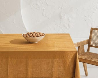 May include: A mustard-colored linen tablecloth covers a wooden table, with a bowl of nuts in the center. A wooden chair with a woven back sits to the right. The background is a textured white wall.