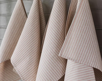 May include: Four beige and red striped kitchen towels hanging from a wooden rack. The towels are folded and have a classic, farmhouse style. The rack is mounted on a gray wooden wall.
