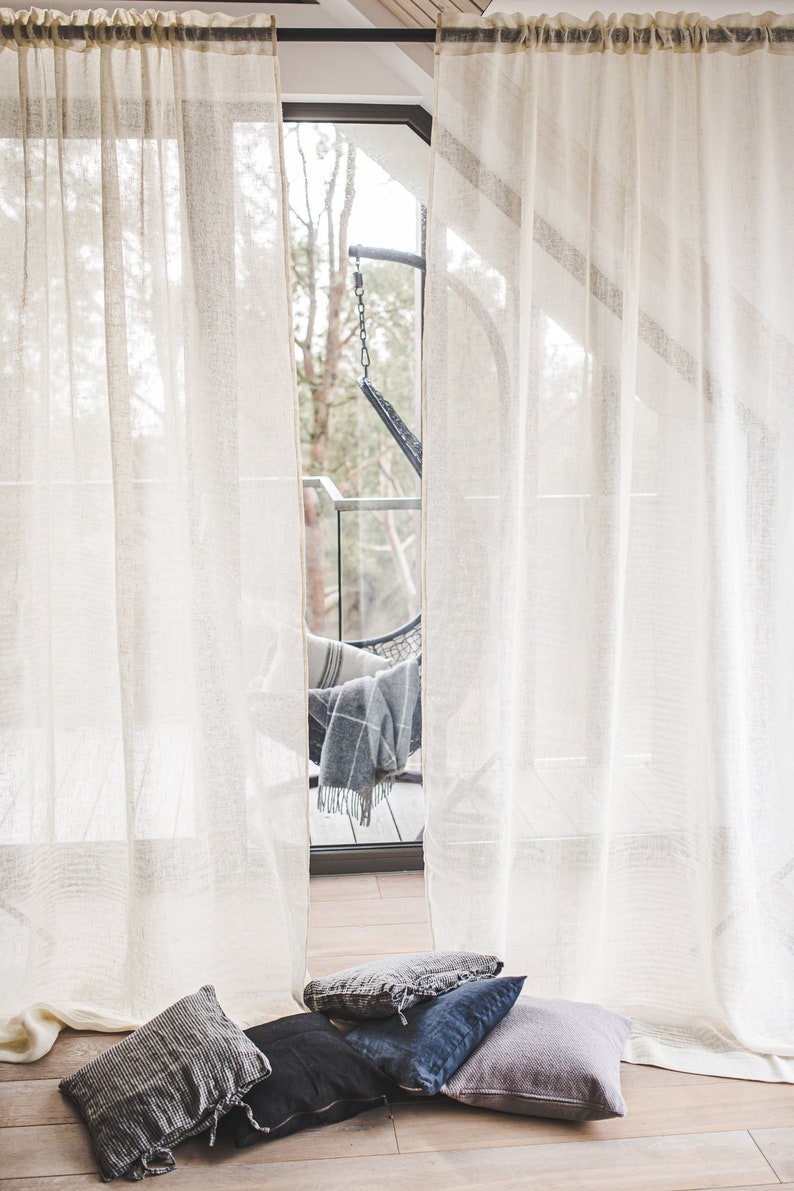 Puede incluir: Dos cortinas semitransparentes de color blanco roto cuelgan de una barra oscura y presentan un sutil patr&oacute;n de rayas horizontales. En el suelo, frente a las cortinas, hay cuatro cojines cuadrados en tonos negro, azul oscuro y gris.