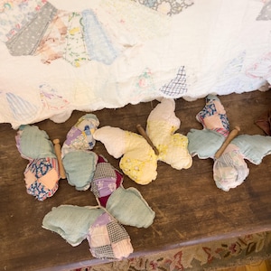 May include: A collection of handmade fabric butterflies with wooden dowel bodies. The wings are made from various patterned fabrics in shades of blue, yellow, purple, and white. They are arranged on a dark wooden surface, with a vintage quilt in the background.