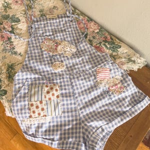 May include: Blue and white gingham romper with floral and lace patches. The romper has adjustable straps and a front pocket with a patchwork design. The garment is made of a lightweight fabric, perfect for warm weather.